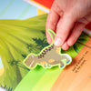 Hand holding a dinosaur sticker over a colorful children's book with a jungle scene.