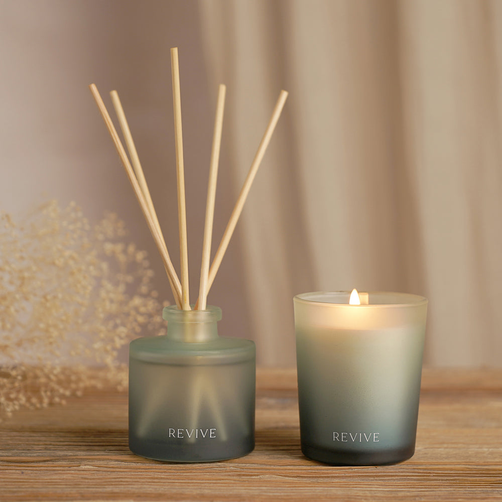 Two 'REVIVE' diffusers, one with sticks and the other a candle, on a wooden surface.