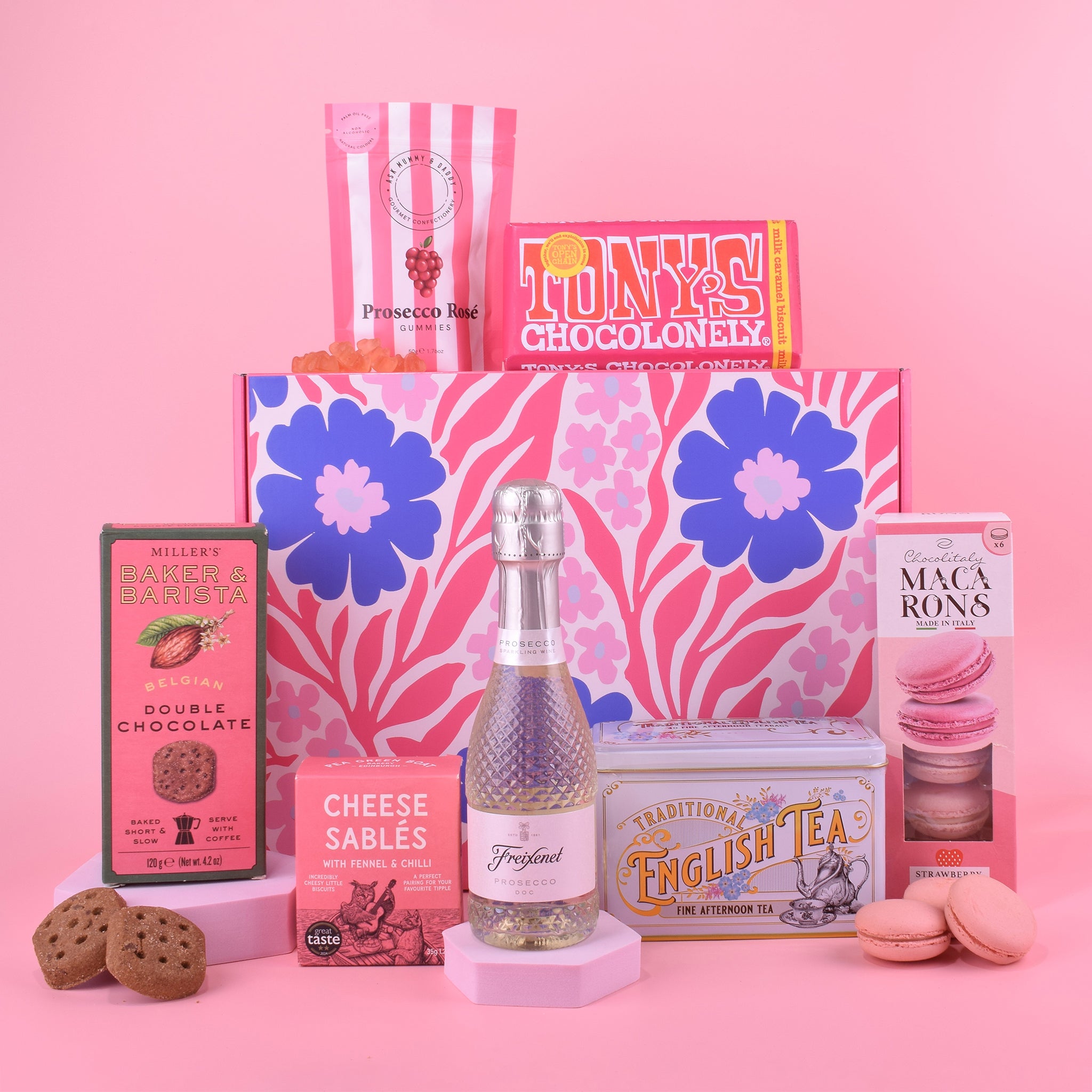 Gourmet gift set with snacks, a bottle of Prosecco, and a box of English tea on a pink background.