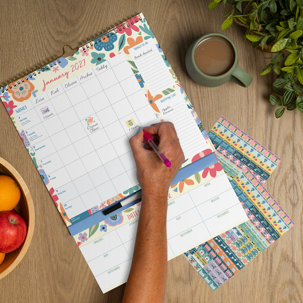 A spiral-bound planner page for January 2021 is laid open on a wooden table, with a hand writing in the date grid using a pink pen. The planner has colorful floral borders and weekly summary bands.