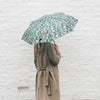 A person seen from behind holding a green umbrella with a pink and white floral pattern, wearing a beige trench coat against a white brick wall.