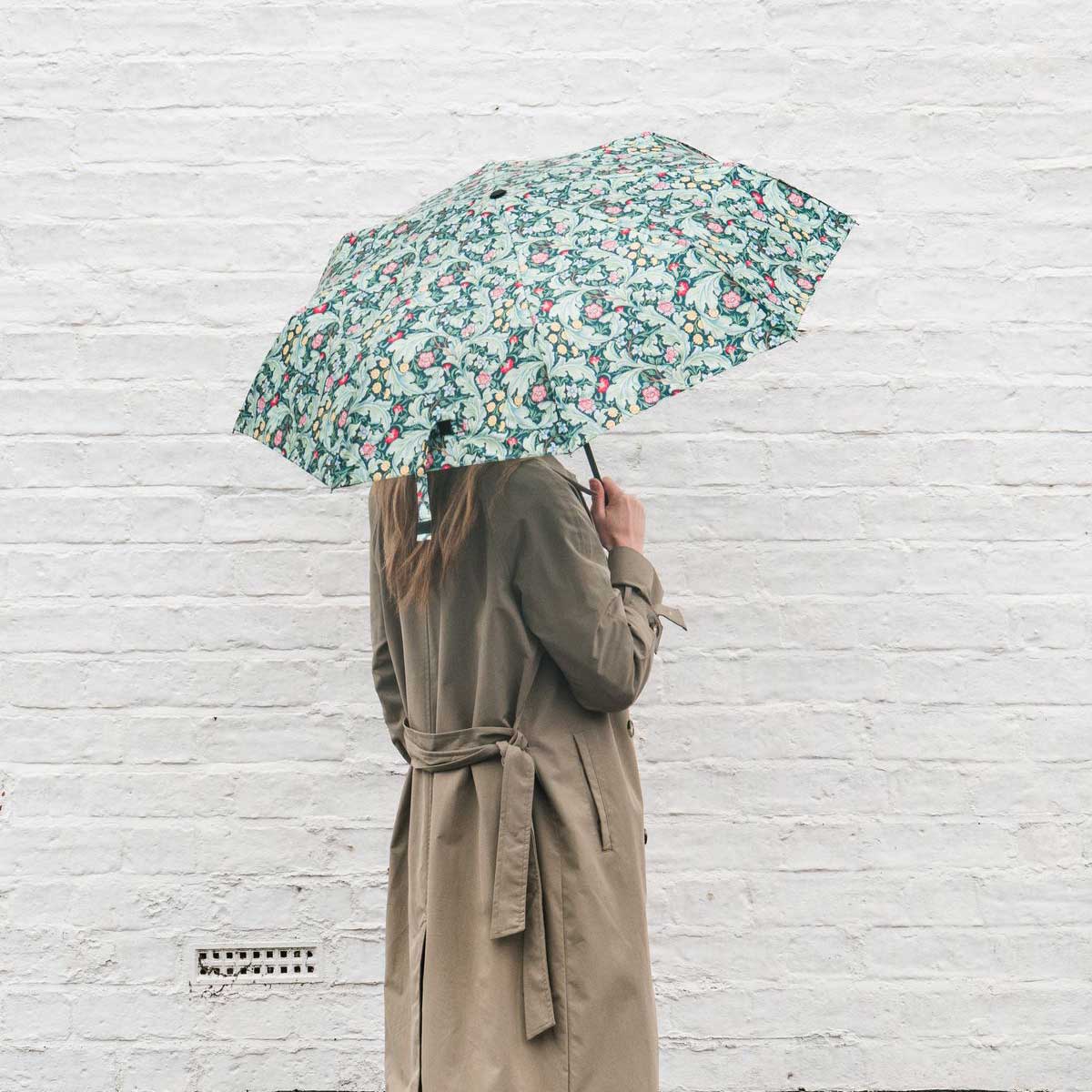 A person seen from behind holding a green umbrella with a pink and white floral pattern, wearing a beige trench coat against a white brick wall.