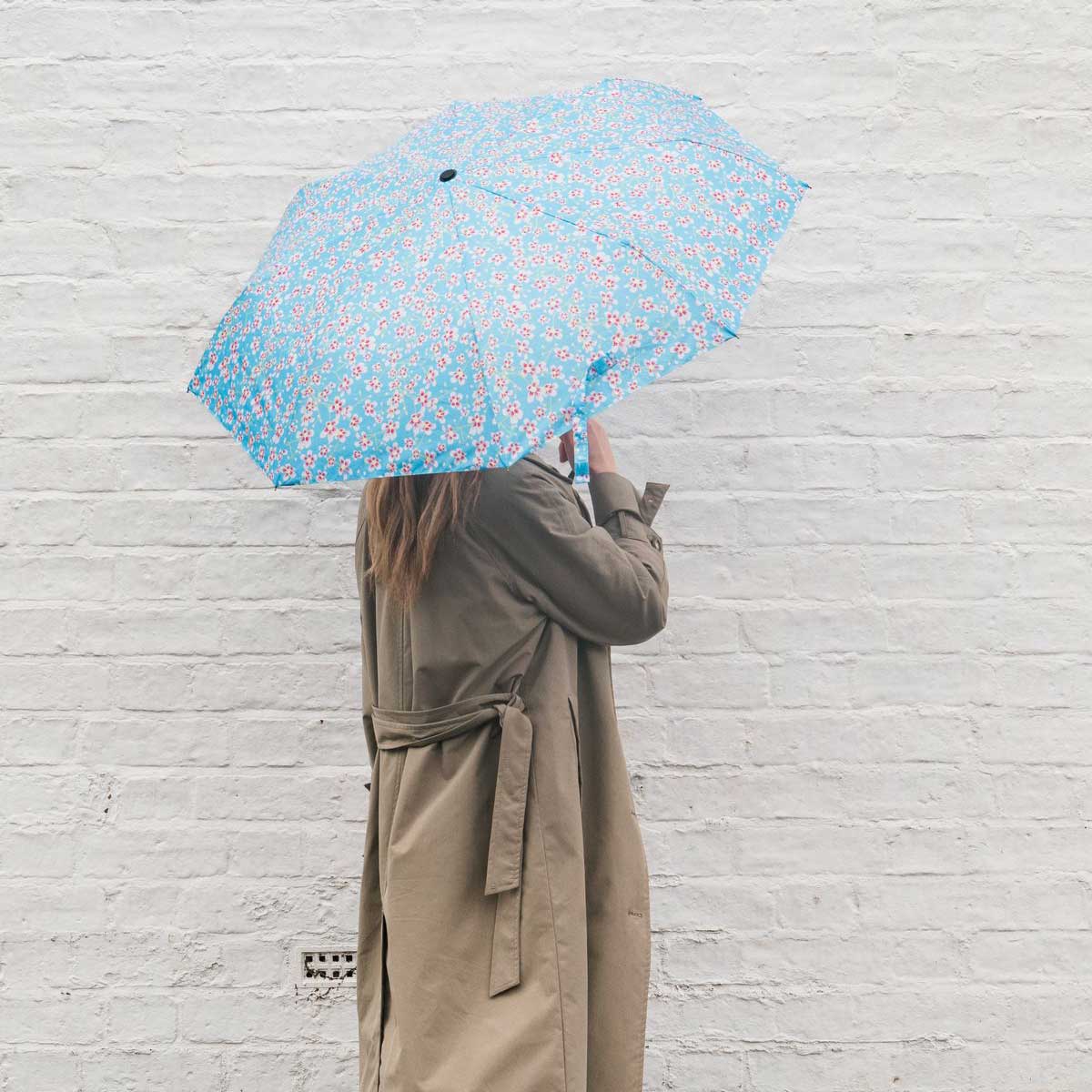 A person in a beige trench coat holds up a light blue umbrella with a pink and white floral pattern against a white brick wall.