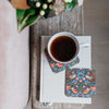 A top-down view of a white ceramic mug filled with black coffee resting on a floral coaster, placed on a white book atop a rustic wooden table. A second matching floral coasters is visible beside the book, with a soft pink upholstered chair and a blurred plant in the background.