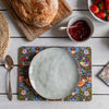 A top-down styled tabletop scene showing a round speckled ceramic plate resting on a colorful floral and bird patterned placemat. Surrounding props include a loaf of bread on a wooden board, a bowl of jam, strawberries, a spoon, and another matching patterned napkin on a white wood plank surface.