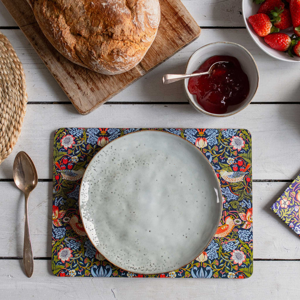 A top-down styled tabletop scene showing a round speckled ceramic plate resting on a colorful floral and bird patterned placemat. Surrounding props include a loaf of bread on a wooden board, a bowl of jam, strawberries, a spoon, and another matching patterned napkin on a white wood plank surface.