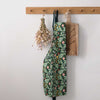 A green floral apron with a botanical pattern hangs from a wooden peg on a white wall, styled with dried flowers and a wooden cutting board.
