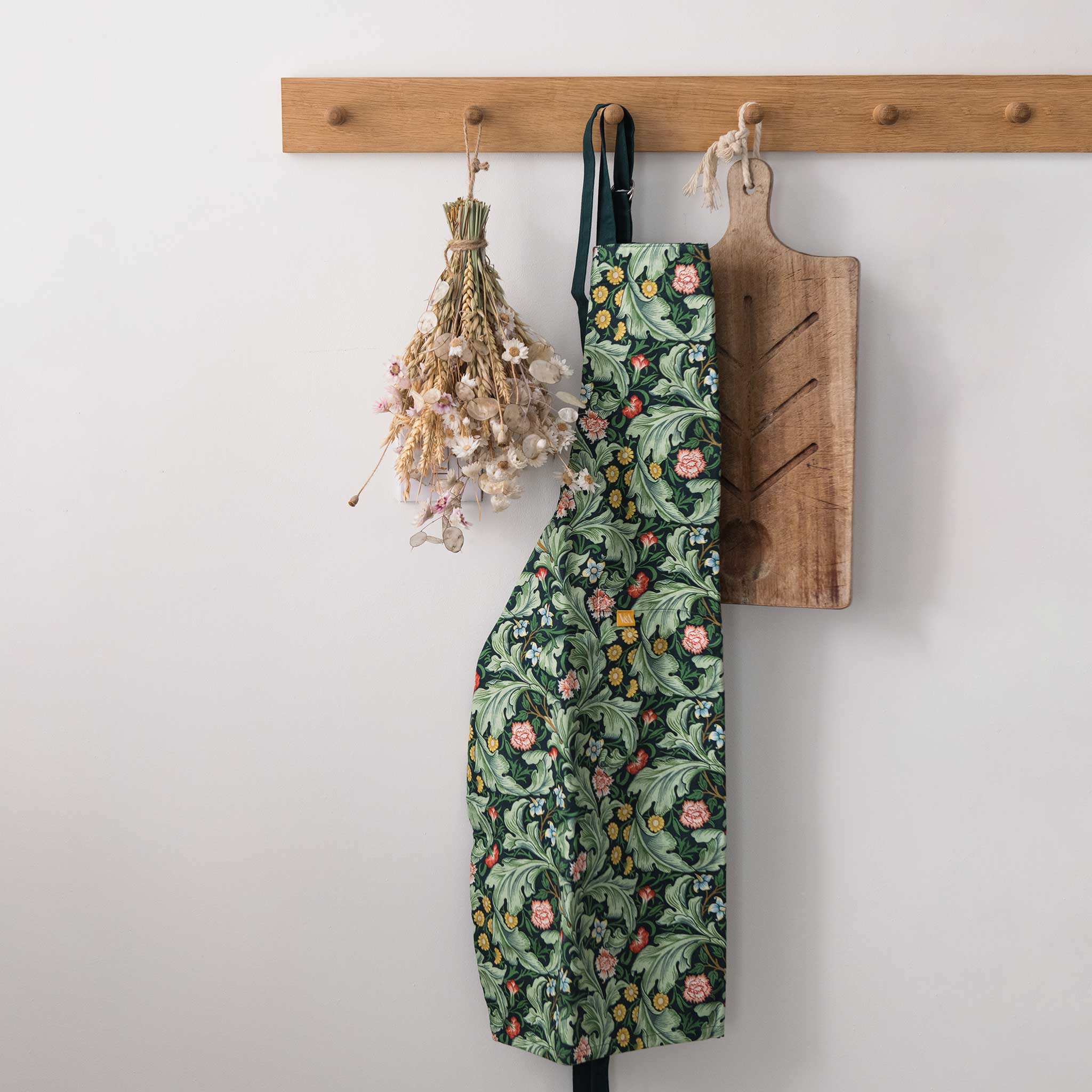 A green floral apron with a botanical pattern hangs from a wooden peg on a white wall, styled with dried flowers and a wooden cutting board.
