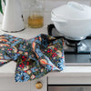 A multicolor floral and bird-patterned kitchen towel draped over a white kitchen counter next to a gas stovetop and a white ceramic pot.