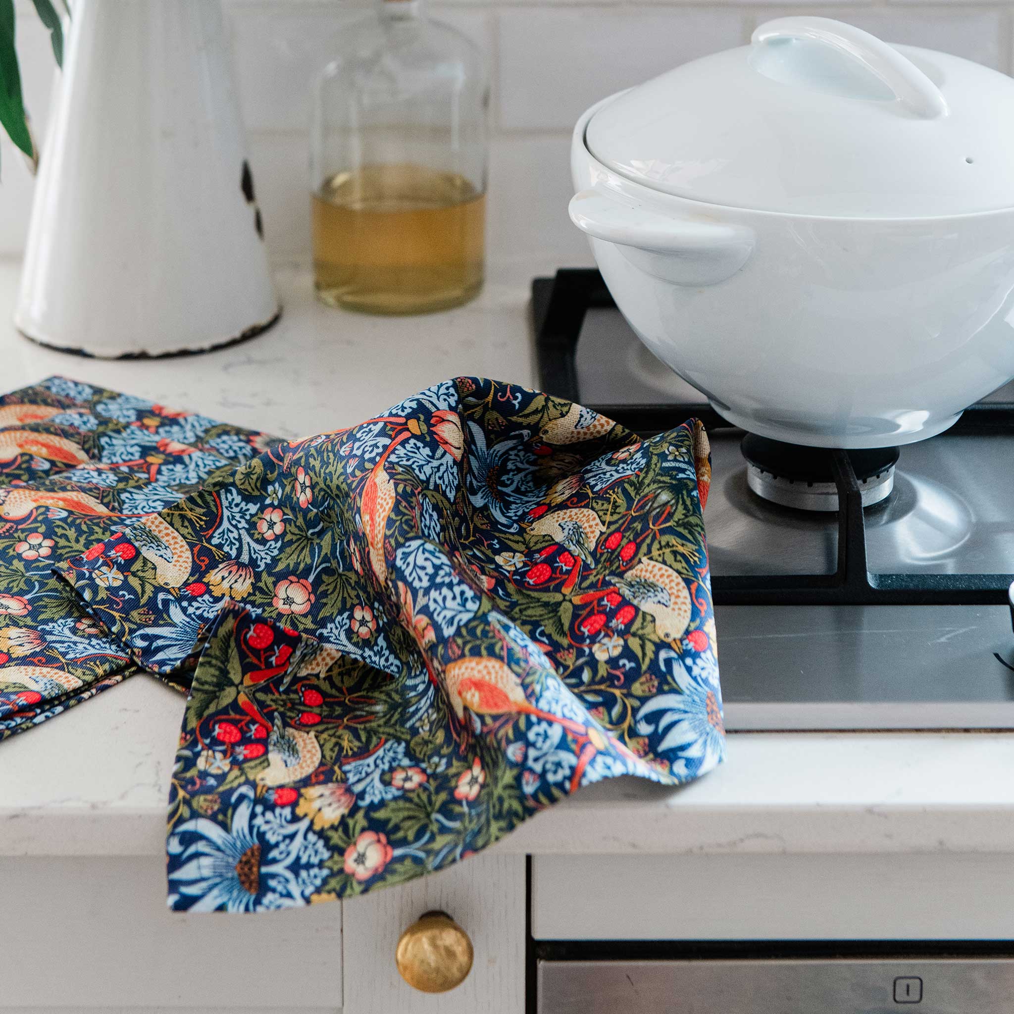 A multicolor floral and bird-patterned kitchen towel draped over a white kitchen counter next to a gas stovetop and a white ceramic pot.