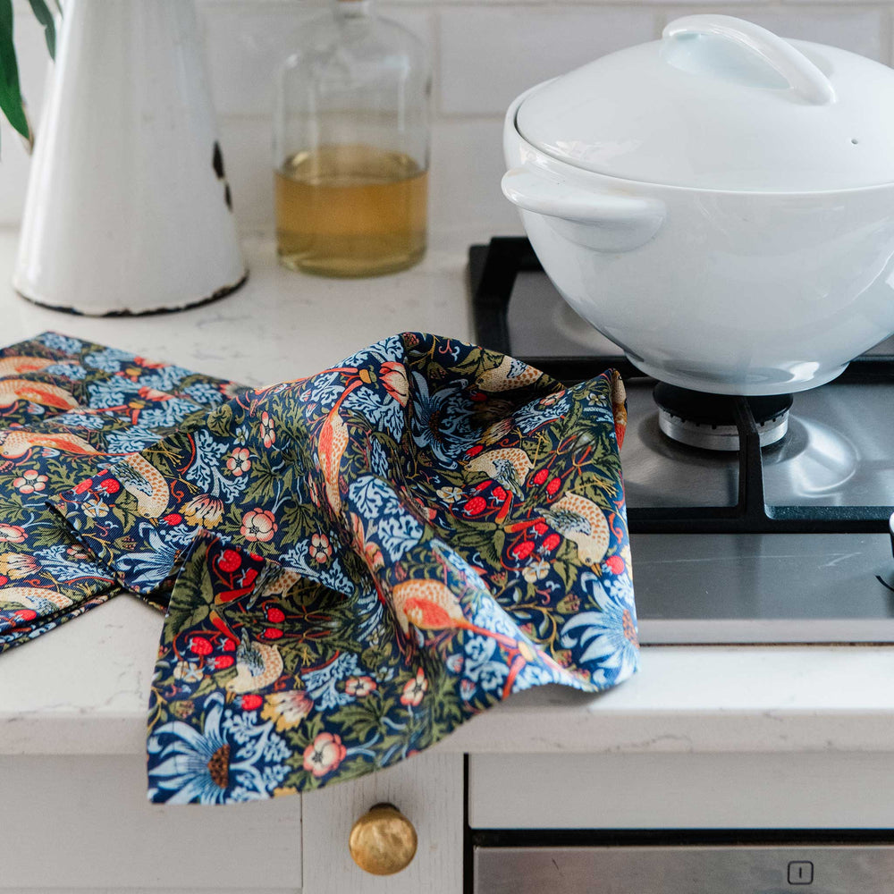 A multicolor floral and bird-patterned kitchen towel draped over a white kitchen counter next to a gas stovetop and a white ceramic pot.