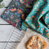 Two patterned kitchen towels displayed on a white wooden surface next to a cooling rack and a pastry. One towel has a dark navy blue background with a detailed bird and floral design in red, orange, and cream.