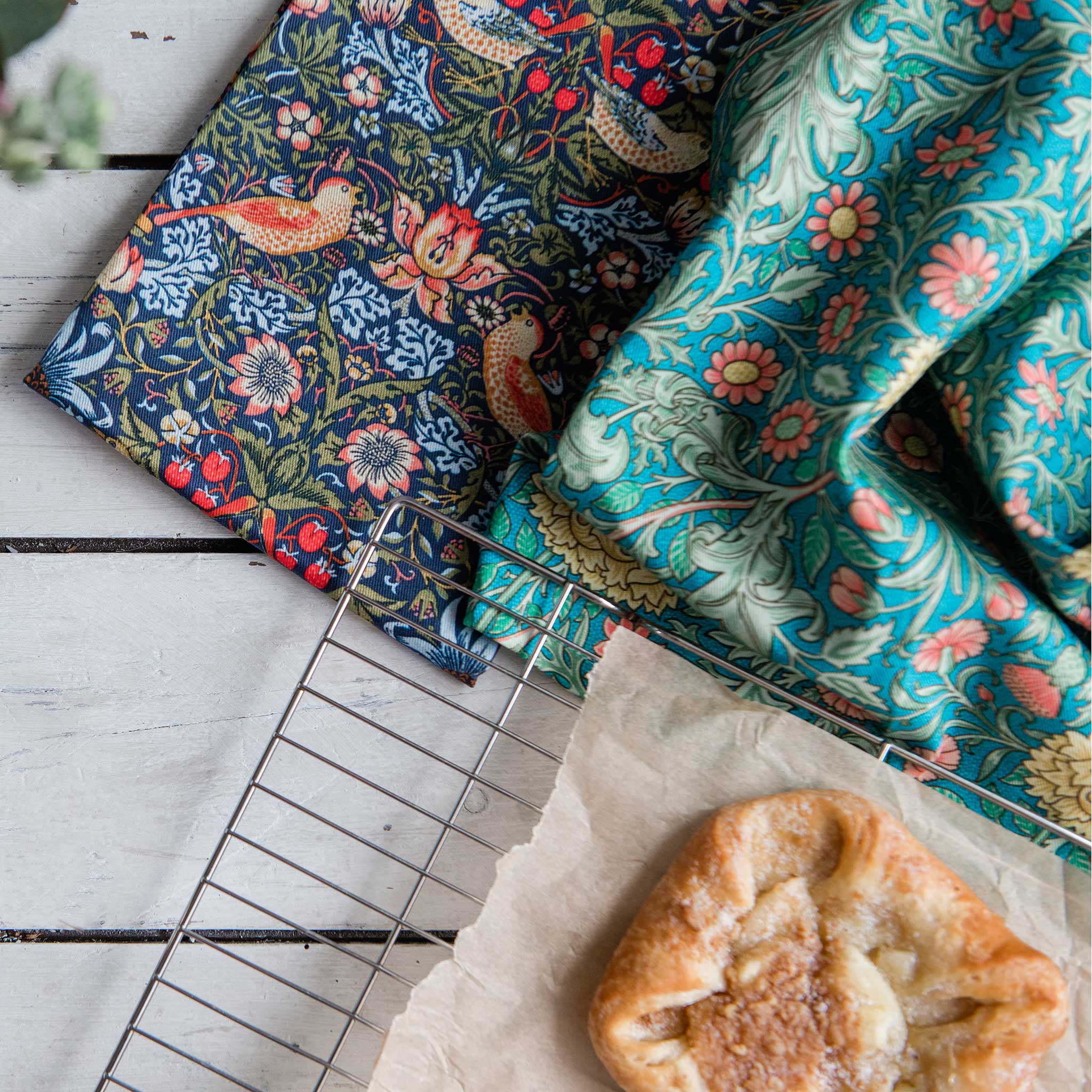 Two patterned kitchen towels displayed on a white wooden surface next to a cooling rack and a pastry. One towel has a dark navy blue background with a detailed bird and floral design in red, orange, and cream.