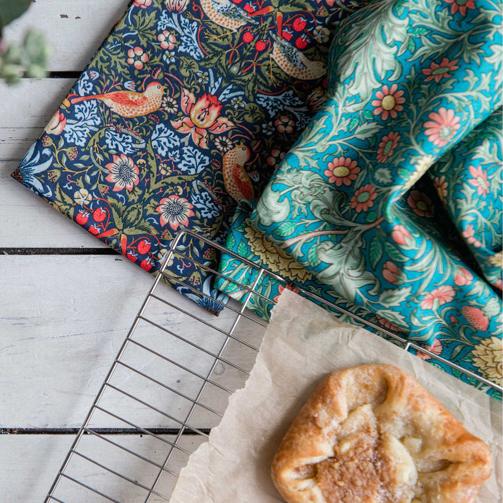 Two patterned kitchen towels displayed on a white wooden surface next to a cooling rack and a pastry. One towel has a dark navy blue background with a detailed bird and floral design in red, orange, and cream.