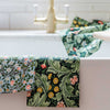 Two folded patterned towels draped over the edge of a white sink, with a matching floral cloth hanging in the background near a gold faucet. The towels feature botanical prints in green, pink, yellow, and white.