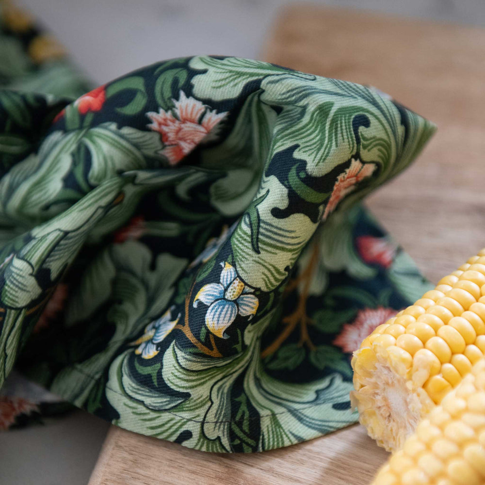 A close-up of a draped fabric with a green and black floral pattern, styled on a wooden surface next to a yellow ear of corn.