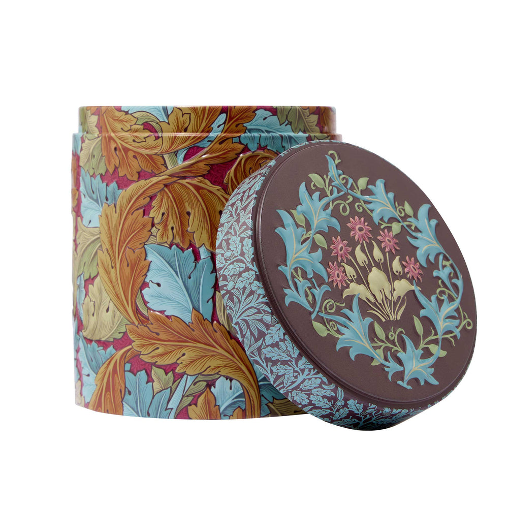 A decorative round metal tin with a matching lid leaning against it, featuring a William Morris-style floral and leaf pattern in blue, gold, brown, and pink on a white background.