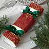 A red Christmas-themed box with gold printed illustrations and text, tied with two green satin ribbons, placed on a white plate with evergreen branches around it.