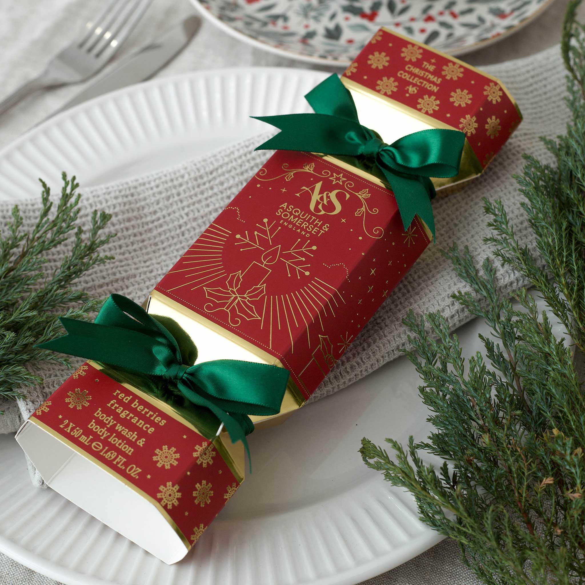 A red Christmas-themed box with gold printed illustrations and text, tied with two green satin ribbons, placed on a white plate with evergreen branches around it.