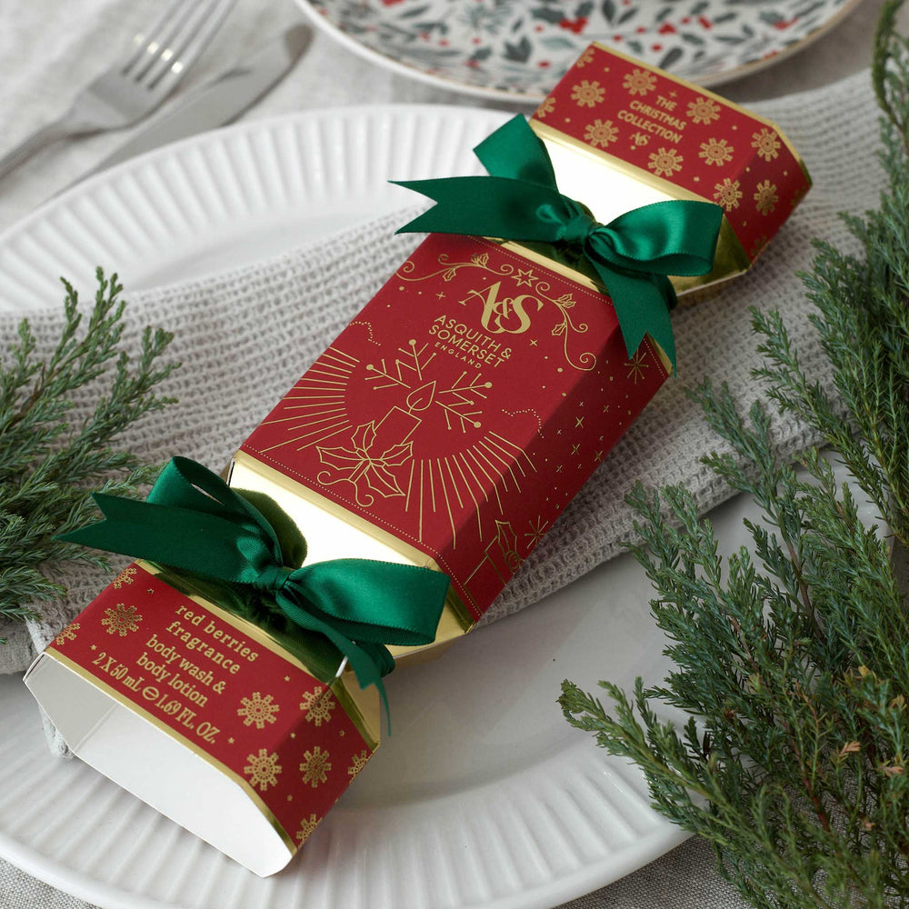 A red Christmas-themed box with gold printed illustrations and text, tied with two green satin ribbons, placed on a white plate with evergreen branches around it.