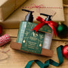 Two ASOUTH & SOMERSET Christmas gift bottles, one green and one white, in a wooden gift box with a green card and red ribbon, styled on a wooden table with wrapped gifts, ornaments, and bakers twine.
