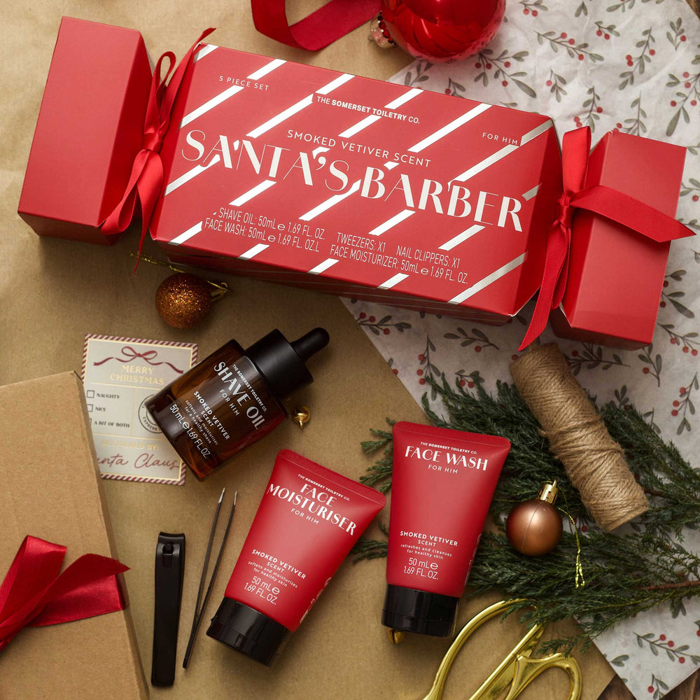 A Christmas-themed grooming gift set from The Grooming Club Co. featuring a red 'Santa's Barber' scented shaving kit box with red gift boxes on either end, arranged on a kraft paper surface with holiday decorations including a bottle of facial oil, face wash, and face washmaster, twine, scissors, and ornaments.