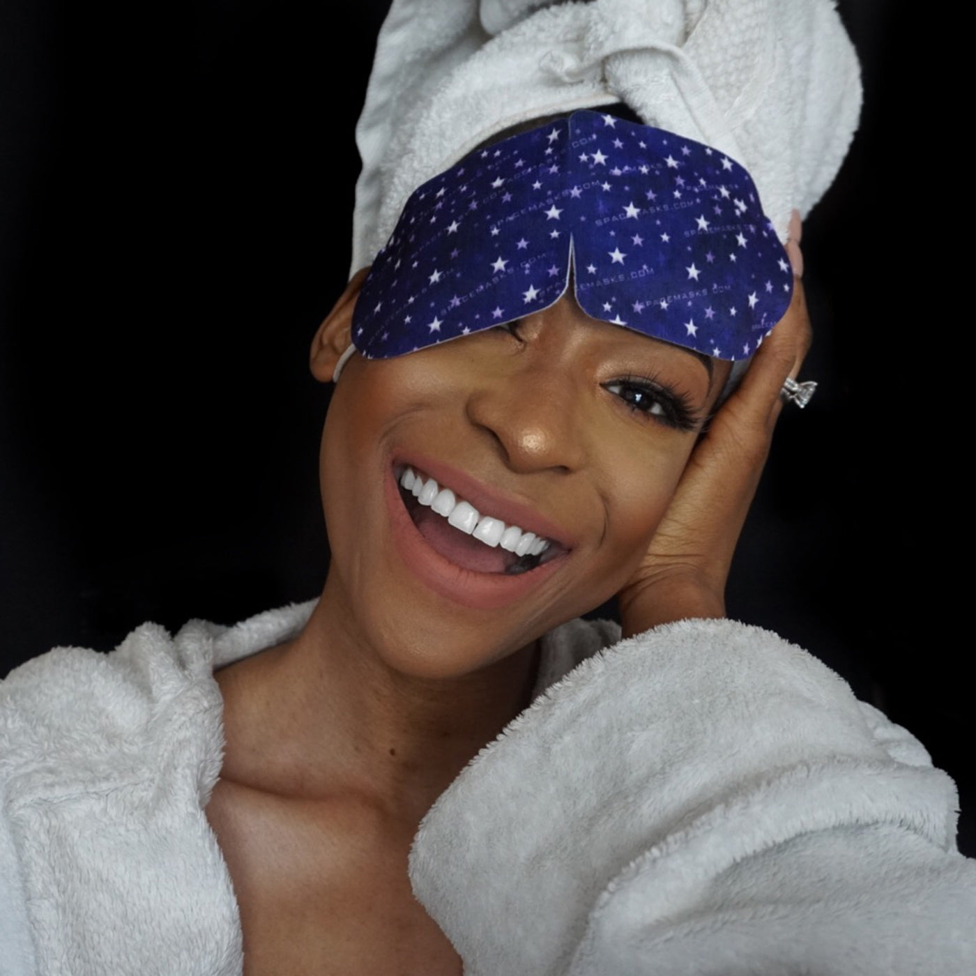 A black woman in white fluffy dressing gown and with her air wrapped in a white towel, smiling while putting on a spacemask